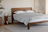 A minimalist bedroom with a wooden bed frame, white bedding, two pillows, a small side table with decor, a large potted plant, a neutral rug, and a light-colored wall with abstract art.