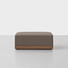 A square, brown upholstered ottoman with a wooden base sits on a light gray floor against a plain white background.
