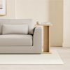 A light beige sofa with a matching rectangular pillow sits next to a wooden side table holding stacked books and a small cup, all on a cream rug in a minimal, softly lit room.