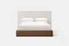 A modern bed with a tall, light beige upholstered headboard, white bedding, and a dark wood base, shown against a plain white background.