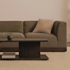 A modern living room with a brown sectional sofa, a light beige cushion, a dark wooden coffee table holding a small vase with greenery, and a book, all set on a light carpet.