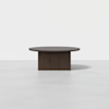 A round, dark brown wooden table with a thick tabletop and a wide rectangular base, placed on a light gray floor against a plain white background.