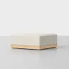 A minimalist, square ottoman with a light beige upholstered cushion and a wooden base, placed on a smooth, light gray surface against a plain white background.
