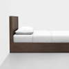 A side view of a modern wooden bed with a tall headboard, white pillow, and white bedding, placed against a plain white wall on a light gray floor.