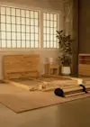 A partially assembled wooden bed frame sits on a beige rug in a modern room with large grid windows, a potted plant, and a matching wooden nightstand and dresser.