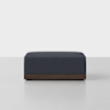 A modern, rectangular ottoman with dark blue upholstery and a wooden base, placed on a light grey floor against a plain white background.