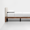 A minimalist wooden bed frame with a beige upholstered headboard, neatly made with white bedding and a pillow, placed against a plain white wall on a light gray floor.