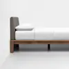 A minimal wooden bed frame with a gray upholstered headboard, white mattress, pillow, and bedding, positioned against a plain white wall in a modern, empty room.