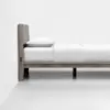 Minimalist wooden bed frame with a padded headboard, dressed in white bedding and a single pillow, set against a plain white wall and floor.