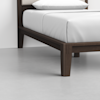 Close-up of the corner of a modern wooden bed frame with dark brown legs, neatly made with white bedding, on a light gray floor against a plain white wall.