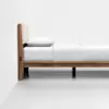 Side view of a minimalist wooden bed with a light-colored upholstered headboard, white pillow, and white bedding, placed against a plain white wall on a light gray floor.