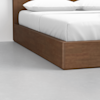 Close-up of the corner of a wooden platform bed with a white mattress and white pillows, positioned on a light gray floor against a plain white wall.