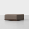A square, brown upholstered ottoman with a low wooden base, set on a light grey floor against a plain white background.