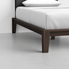 A close-up view of a modern bed with a dark wooden frame, white bedding, and a pillow, set on a light grey floor against a plain white wall.