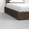 A close-up view of the corner of a modern bed with a dark wooden frame, white bedding, and a pillow, set on a light gray floor against a white wall.