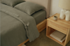 A neatly made bed with sage green bedding and two pillows is beside a light wooden nightstand holding a small ceramic cup, a lamp, and two closed books on the lower shelf.