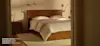A neatly made double bed with white linens, a light beige blanket, and a wooden headboard sits in a softly lit bedroom with beige walls, two wall-mounted lamps, and a nightstand with a plant.