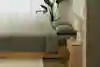 A minimalist bedroom with a wooden bed frame, green bedding, a wooden nightstand with a lamp and cup, a potted plant by the window, white curtains, and neutral-colored walls and floor.