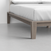 Close-up of the corner of a wooden bed frame with a light brown finish, white mattress, and white bedding, set on a light gray floor in a minimal, modern room.