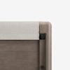 Close-up of the top corner of a wooden bed frame with a light beige upholstered headboard panel, featuring smooth rounded edges and visible assembly hardware.