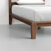 A close-up view of a wooden bed frame with a white mattress and pillow, set against a light gray floor and white wall. The bed frame features clean lines and a modern design.