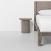 A minimalist bedroom scene featuring a wooden bed frame with white bedding and a matching wooden nightstand on a light gray floor against a white wall.