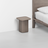 A minimalist wooden nightstand with a rounded square top, placed next to a wooden bed with white bedding in a bright, modern room with white walls and floor.