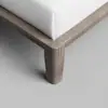 Close-up view of a modern wooden bed frame corner with a light brown finish and a white mattress on top, photographed against a light gray floor. The bed frame has clean lines and a sturdy, geometric leg.