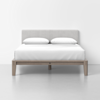 A modern bed with a light gray upholstered headboard, white bedding, two white pillows, and a minimalist wooden frame, set against a plain white background.