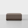 A modern, rectangular brown ottoman with a fabric cushion top and a dark wooden base, placed on a light gray floor against a white background.