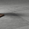 A section of gray carpet flooring with visible seams and a small corner of a brown rectangular object in the lower left corner.