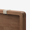 A close-up of the corner of a wooden object, possibly a table or panel, with a light-colored rectangular clip attached to the edge. The wood has a smooth finish and rounded corners.