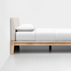 Side view of a minimalist bed with a light wood frame, beige upholstered headboard, and neatly made white bedding and pillow, against a plain white wall and floor.
