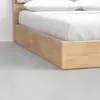 Close-up of a wooden bed frame with a light wood finish, featuring crisp white bedding and pillows, set on a smooth, light gray floor against a white wall.