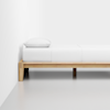 A minimalist wooden bed frame with a white mattress, white bedding, and a single white pillow, placed against a plain white wall on a light gray floor.
