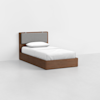 A wooden single bed with a gray cushioned headboard is placed in a minimalistic, empty white room. The bed has white bedding and a single pillow.