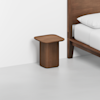 A modern wooden nightstand with rounded edges sits next to a wooden bed frame with a white mattress and pillow, against a plain white wall and light-colored floor.