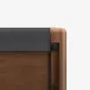 Close-up of the corner of a wooden bed frame with a dark gray fabric-covered headboard, showcasing smooth, rounded wood edges and visible metal fasteners.