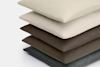 A stack of four rectangular pillows in muted, neutral colors—beige, light gray, brown, and dark gray—arranged on a plain white background.