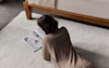 A person with dark hair, wearing a beige top, is lying on a light-colored rug and reading a magazine with large bold letters, next to a wooden bed.