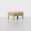 A minimalist wooden stool with a light beige upholstered cushion, placed on a light grey surface against a plain white background.