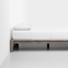A minimalist bed with a light wood frame, white bedding, and a single white pillow, positioned against a white wall on a light-colored floor.