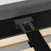 Close-up of a bed frame assembly showing a black metal support bracket attached to a headboard. Wooden slats and a black fabric strap are visible in the foreground.