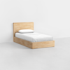 A single wooden bed with a headboard, made of light-colored wood, is placed against a plain white wall. The bed is neatly made with white sheets and a white pillow. The floor is smooth and light gray.