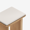 A close-up of a wooden stool with a light-colored, cushioned fabric seat on top, set against a plain white background.