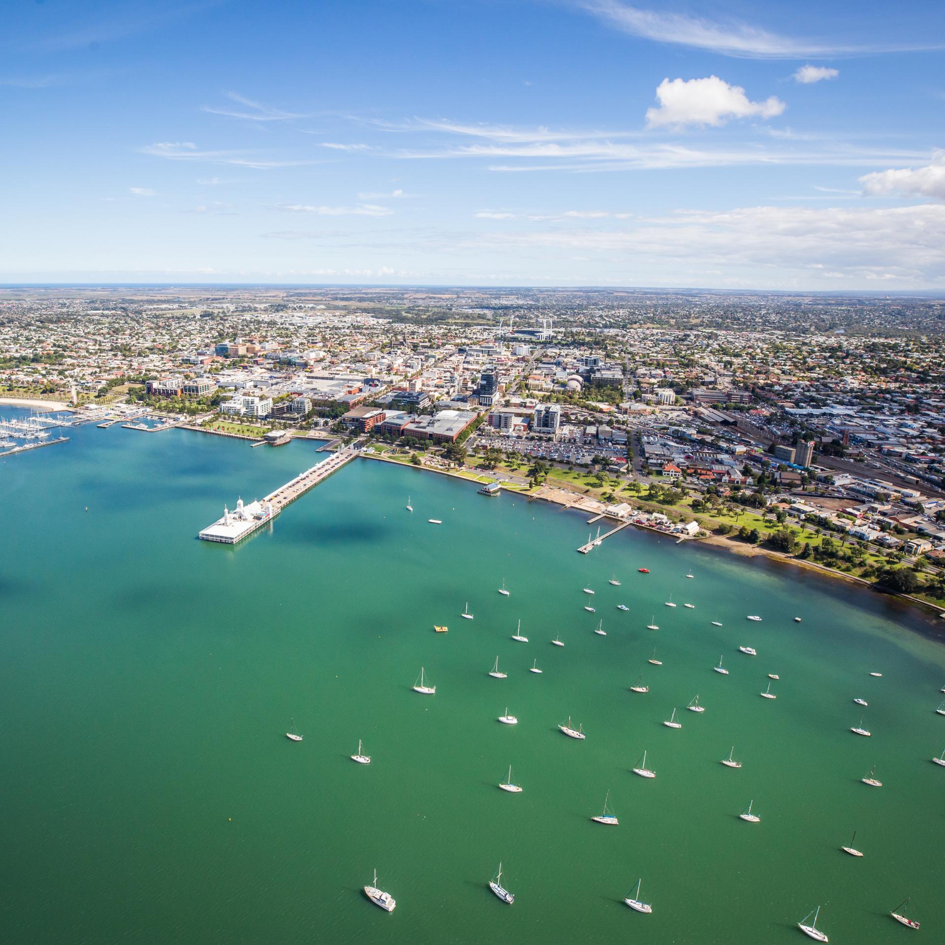 Bird's view of the waterfront of Geelong on a bright day with blue skys. Boats are scattered throughout the greenish water. 