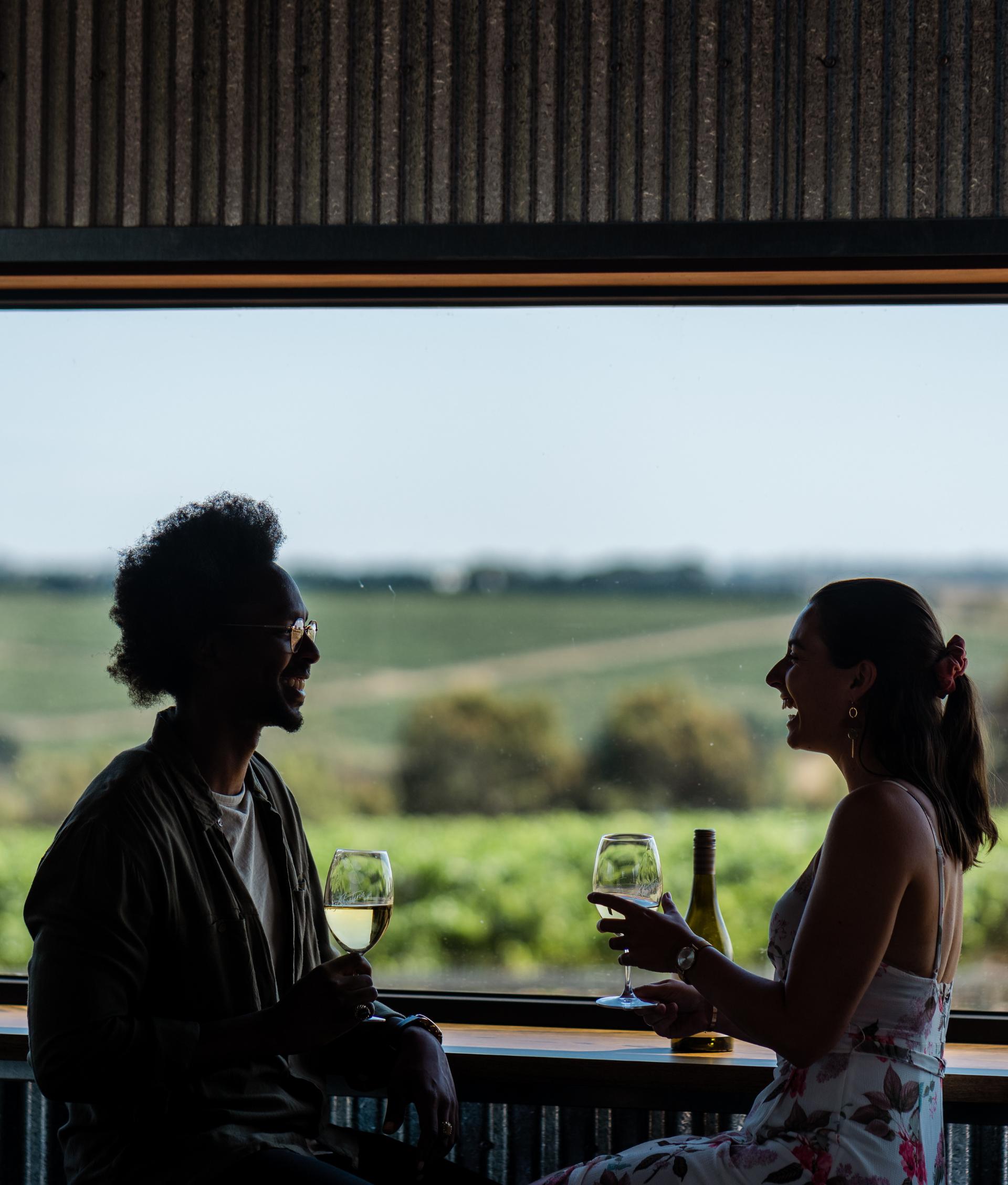 Two people sitting in front of a window drinking wine, laughing with one another. 