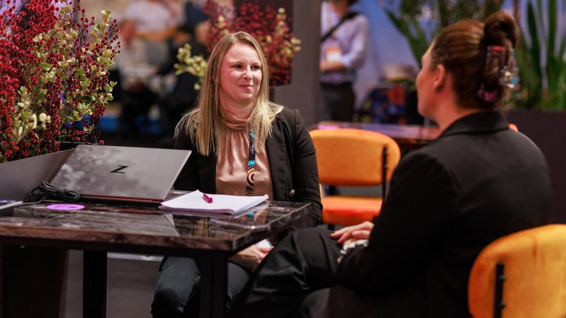 Two woman sitting at a table in conversation with a notebook and laptop on the table in front of one of them. 