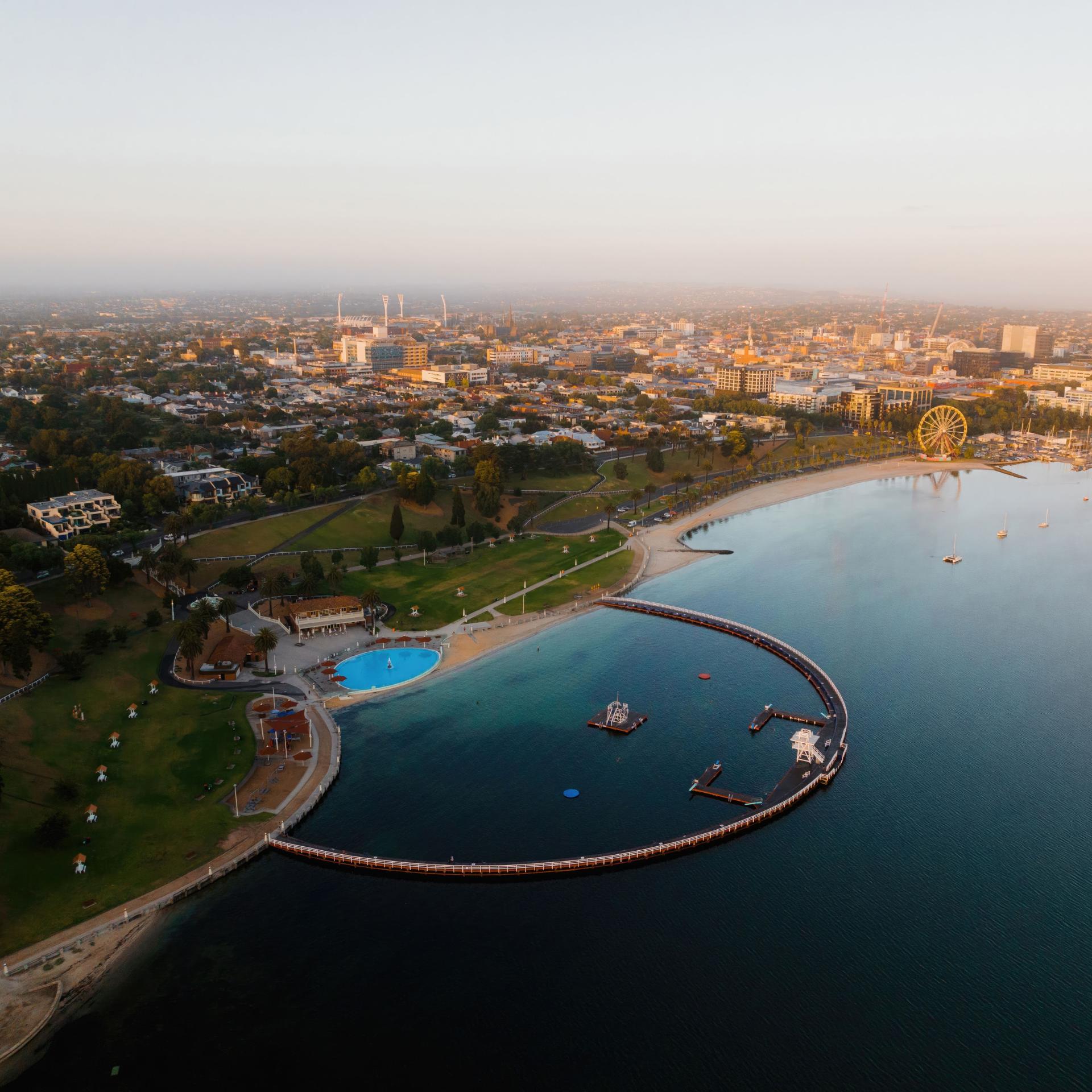 Bird's view of the Geelong waterfront. A large arched walking pier hooks through the water and a fountain faces it on the beach front. A ferris wheel can be seen in the background. 