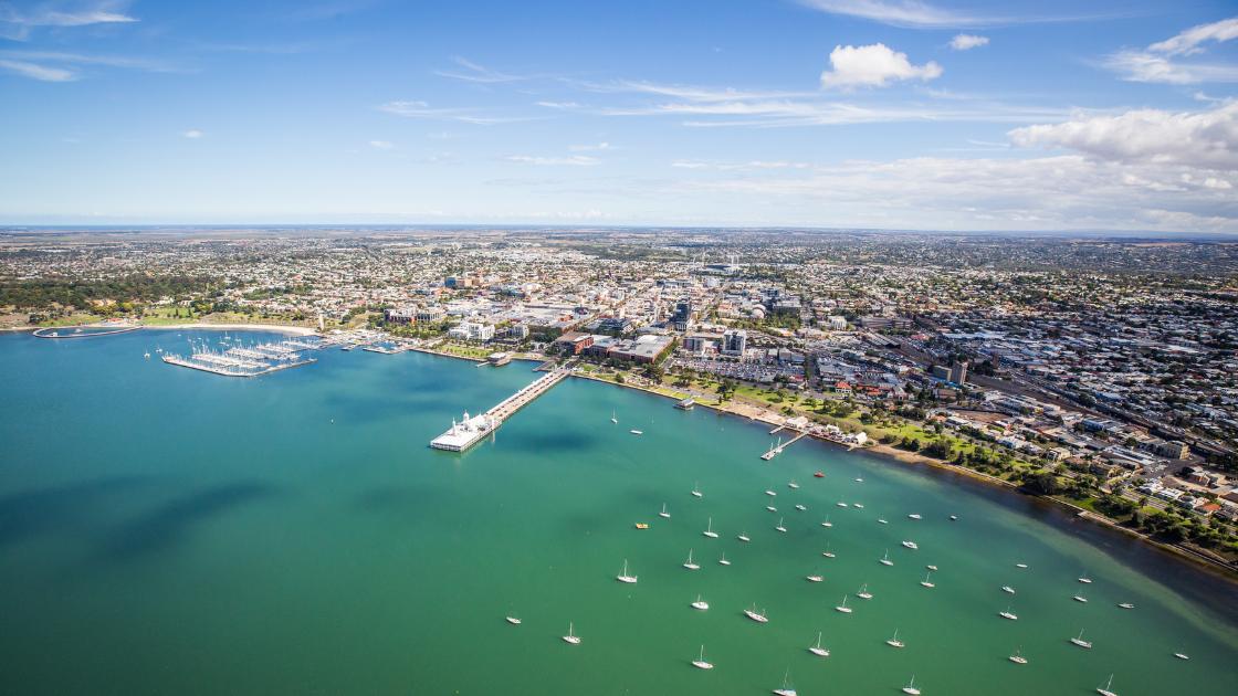 Bird's view of the shoreline of Geelong on a bright day with blue skies. Boats are scattered throughout the greenish water.